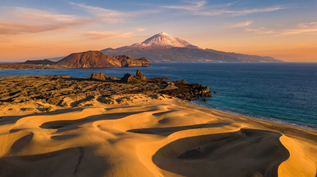 Dunes de sable dorées au premier plan avec le volcan enneigé du Teide en arrière-plan sous un ciel de coucher de soleil.