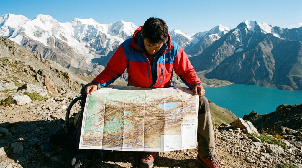 Carte du Kirghizistan : ton guide pour explorer les sommets Un randonneur en veste rouge et bleue consulte une carte du Kirghizistan devant des sommets enneigés et un lac turquoise.